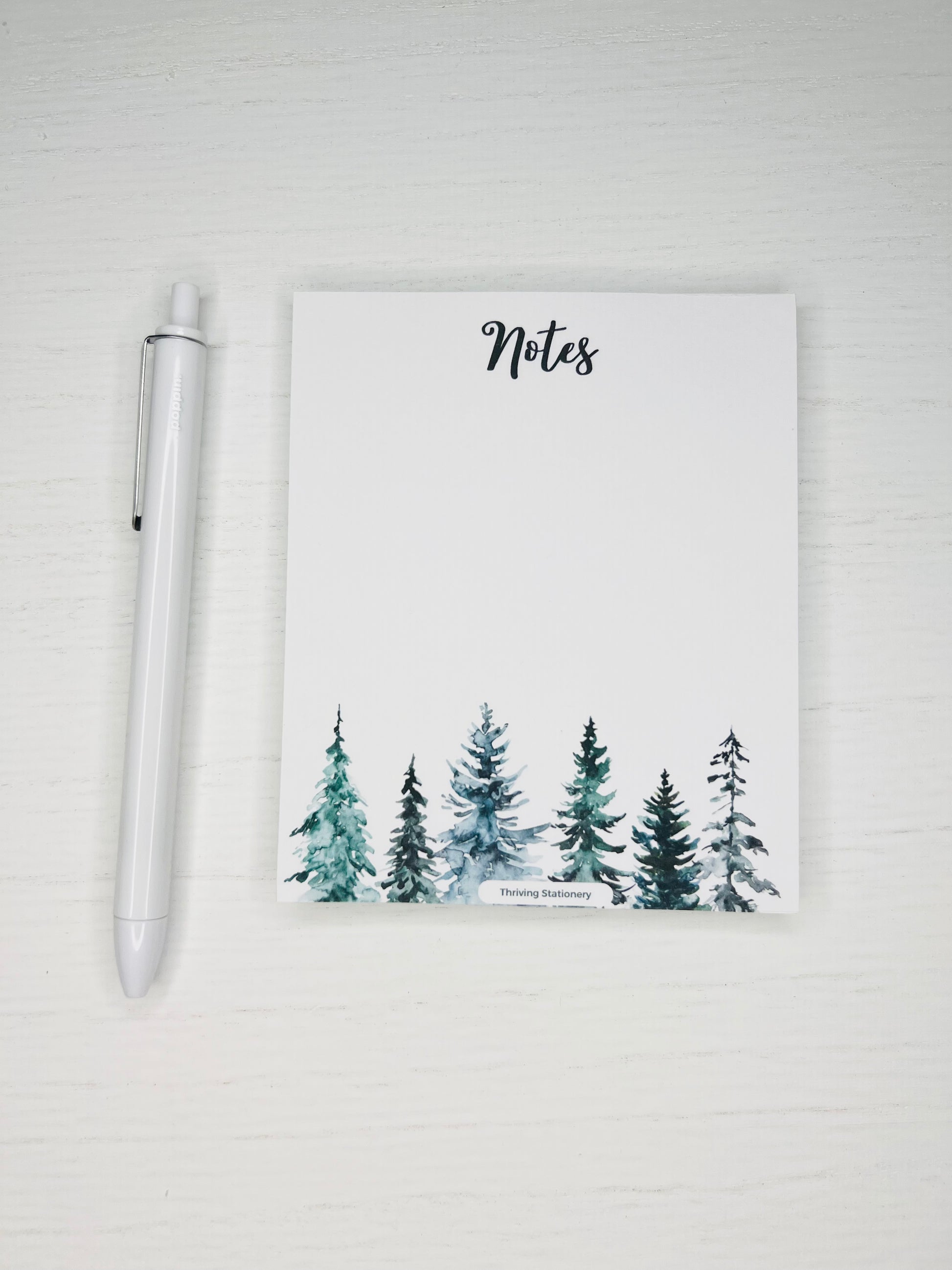 A blank notepad with a winter tree design, accompanied by a white pen, placed on a white surface.